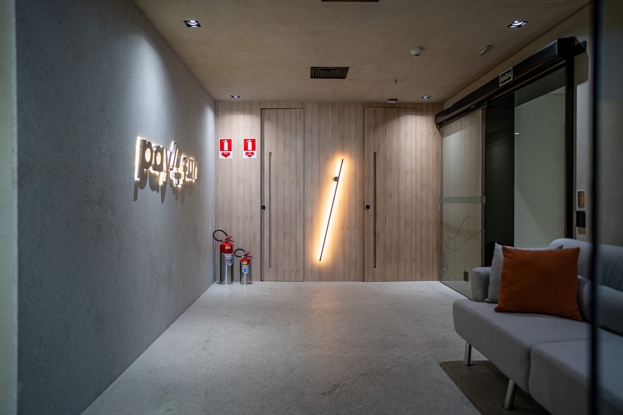 Recepção da Pay4Fun, com logo iluminado sobre revestimento em pedra e portas de madeira